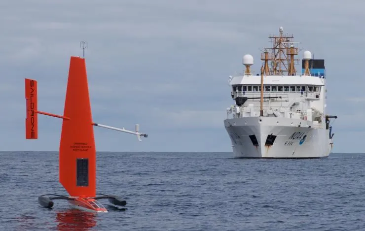 An orange Saildrone Explorer sailing near a NOAA research ship.